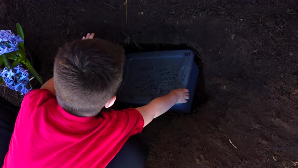 A young boy placing the plastic case of the body of a dead family pet in a recently dug hole in a fa
