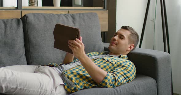 Young Man Is Lying on a Couch and Uses His Tablet