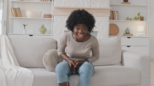 Excited Emotional Young Adult African American Lady Sit Alone on Couch at Home Holding Remote