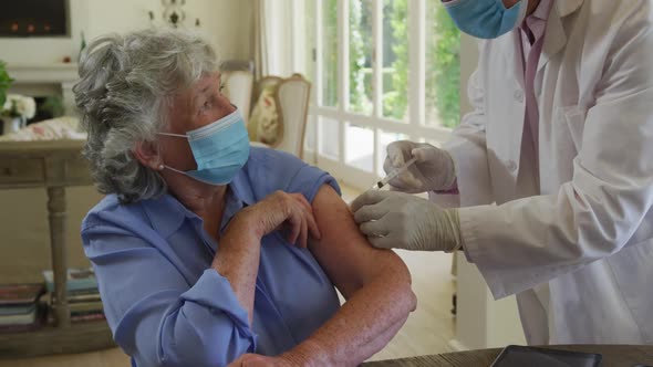 Caucasian senior male doctor wearing face mask injecting covid-19 vaccine into senior woman at home