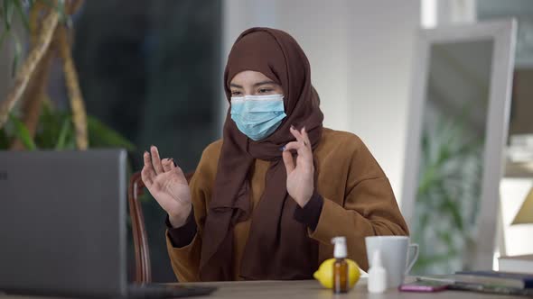 Medium Shot of Young Muslim Woman in Hijab and Covid Face Mask Conferencing in Home Office Sneezing