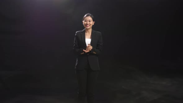 Smiling Asian Speaker Woman Clapping Her Hands And Looking Around While Standing In The Black Studio