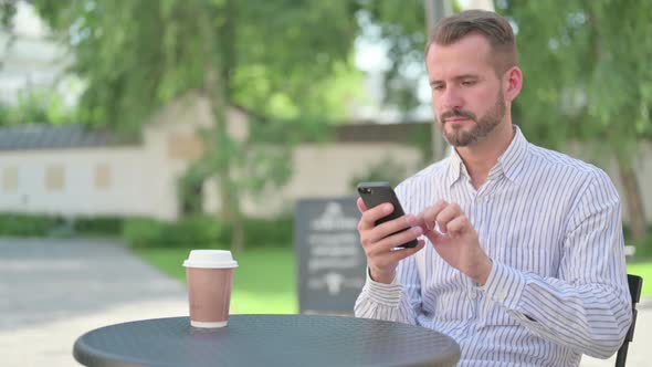 Middle Aged Man Browsing Internet on Smartphone in Outdoor Cafe