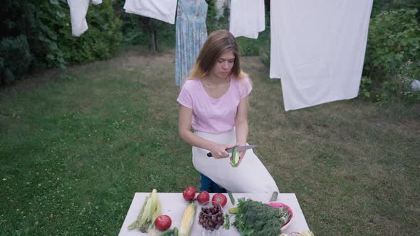 High Angle View of Focused Young Housewife Peeling Cucumber in Slow Motion Sitting on Backyard with