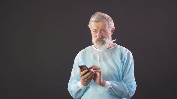 Portrait of Old Surgeon. Old Surgeon Uses Smartphone on Dark Background.