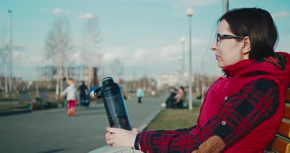 Woman in Glasses with Reusable Plastic Bottle Sitting in Public Park on Bench