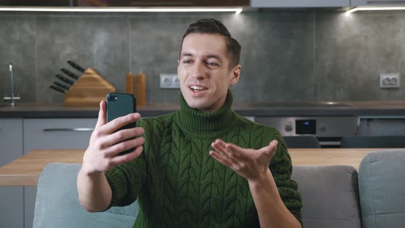 Smiling Man in Sweater with Smartphone Making Video Call While Sitting on Couch in Living Room