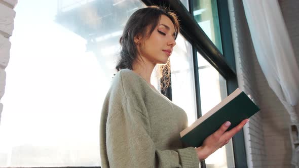 Beautiful Woman Sits Near Window and Reads a Book on Background of Sunset