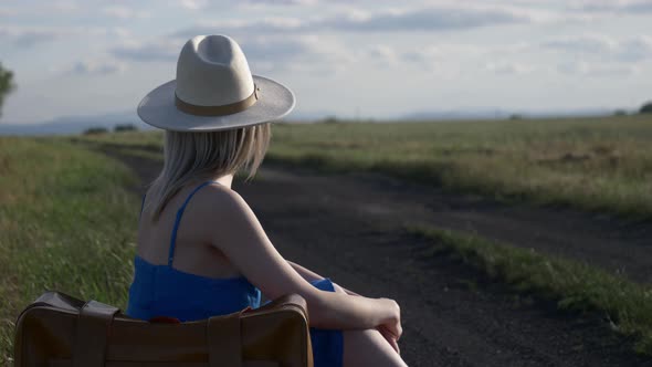 Girl in blue dress with suitcase sit on country road in summer.