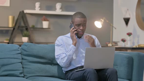 Angry African Man Talking on Smartphone While Using Laptop on Sofa