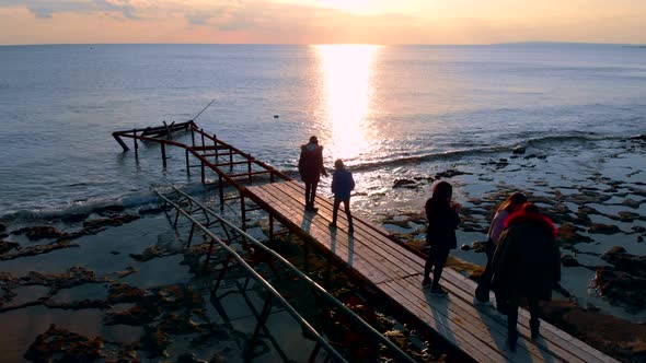 People Are Walking Over Old Broken Wooden Pier and Viewing Sunset Over Sea in Cold Day, Aerial View