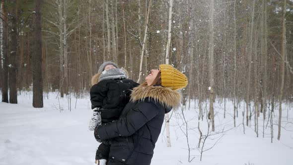 Young Mom with Little Son in Her Hands is Looking at Snowfall in Winter Forest