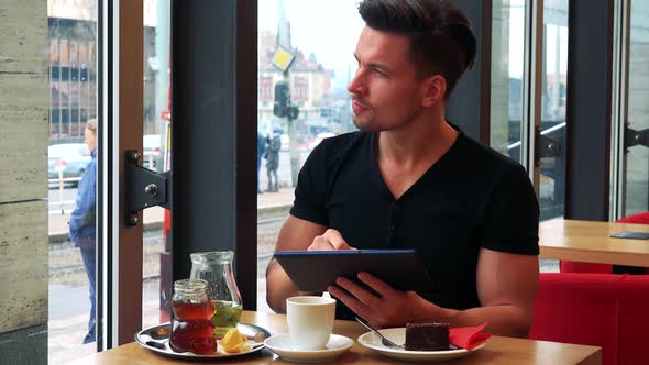 A Young Handsome Man Sits at a Table with Meal in a Cafe and Works on a Tablet