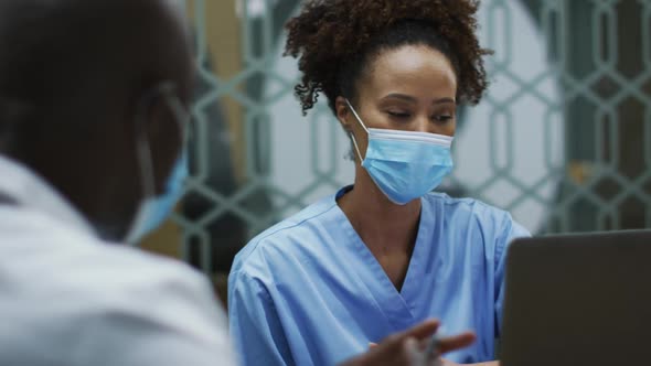 Diverse female and male doctors wearing face masks discussing using laptop in office