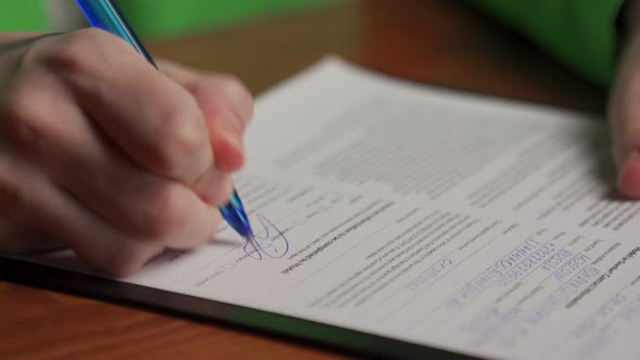 Woman Signs Document with Pen Sitting at Wooden Table