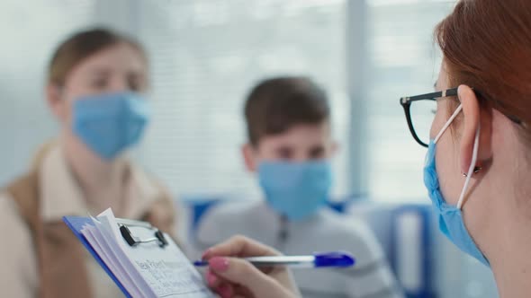 Family Medical Doctor in Mask and Glasses Writes on Clipboard During Patients Consultation in
