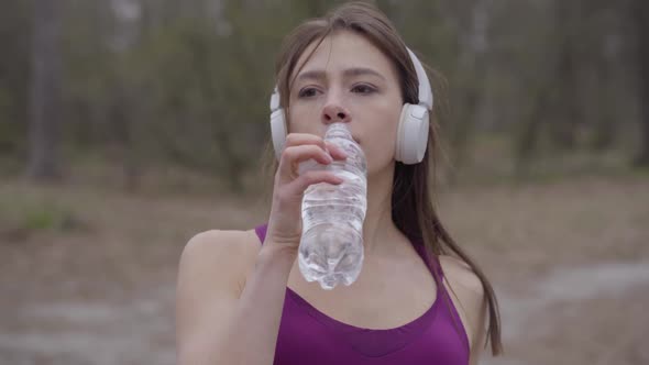 Portrait of Cheerful Young Caucasian Sportswoman in Headphones Drinking Water During Workout Break