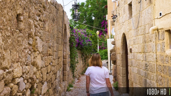 Following young woman walk down narrow cobblestone street with stone walls and houses