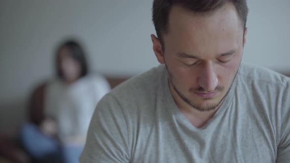 Close-up Portrait of Crying Man on the Foreground and His Shouting Angry Wife Sitting on the Sofa