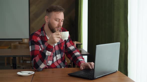 Young Caucasian Man Using Laptop Computer Sitting at Table in Home Office Drinking Coffee Reading