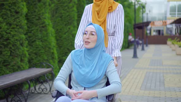 Young Muslim Disabled in a Traditional Scarf in a Wheelchair Communicates with a Muslim Friend 