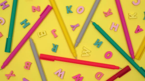 colored ballpoint pens and alphabet letters randomly lie on rotating yellow background. flat lay. 4k