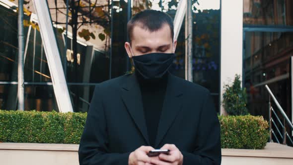 Young Caucasian Businessman in Medical Mask Walking the Street and Texting Message on Mobile Phone