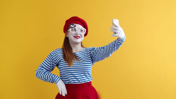 Mime woman takes a selfie contorting her face and smiling, on an orange background.