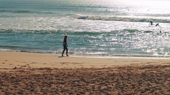 Female surfer walking down the Atlantic Ocean beach at sunset.