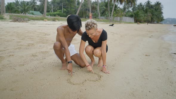 Interracial Couple with Age Differences Drawing a Heart Shape on Sandy Beach and Kissing During a