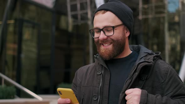 Crop View of Man in Glasses Looking at Phone Screen and Smiling While Walking at Street. Bearded Guy