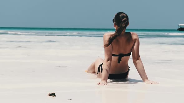 Young Woman Sunbathes on a Paradise Sandy Beach Sits in Black Bikini Near Ocean