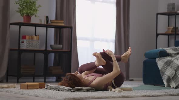Woman is Doing Physical Exercise for Flexibility Lying on Floor in Living Room Holding Legs in
