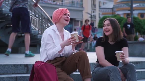 Two Young Women Talking and Laughing Sitting on the Granite Stairs and Drinking Coffee
