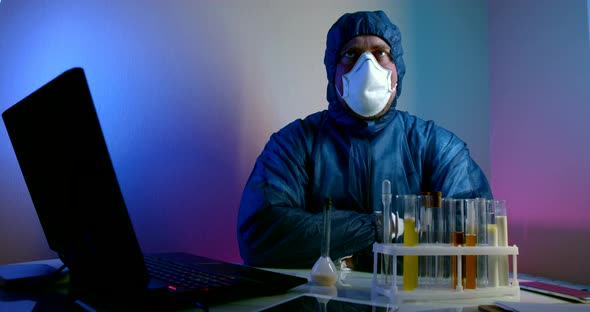 a Man in Blue Protective Clothing with a Hood, Gloves and a Respirator Is Sitting at a Table with