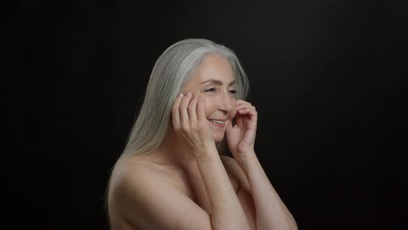 Beautiful Grey Haired Senior Lady with Bare Shoulders Doing Facial Self Massage Black Background