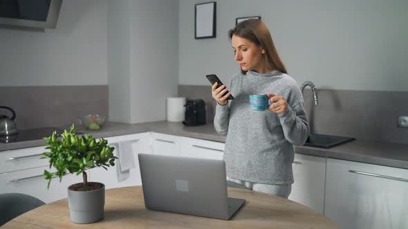 Woman with Smartphone in Hand Standing in Home Cozy Kitchen Use Laptop Do Freelance Work Looking