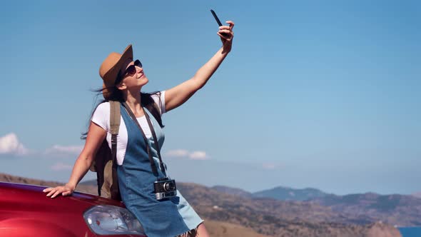 Smiling Travel Woman with Waving Hair Posing on Car Bonnet Taking Selfie Using Smartphone