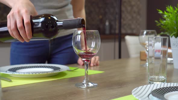 Women's hands pour red wine into a glass on the dining table in the kitchen.
