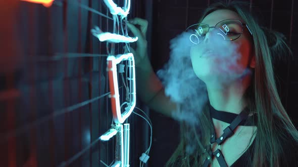 Sexy Model Caucasian Girl in Neon Blowing the Smoke of an Electronic Cigarette or Vape