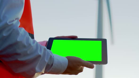 The hands of a man in a shirt and orange vest hold a digital tablet with a green screen, against the