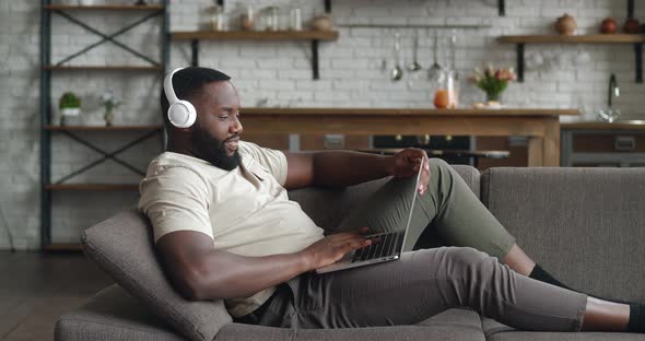 Side View African American Man in Headphones Leaning on Sofa Working Remotely on Laptop From Home