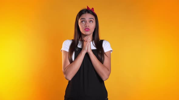 Cute European Young Girl in Black Overall Praying Over Yellow Background. 