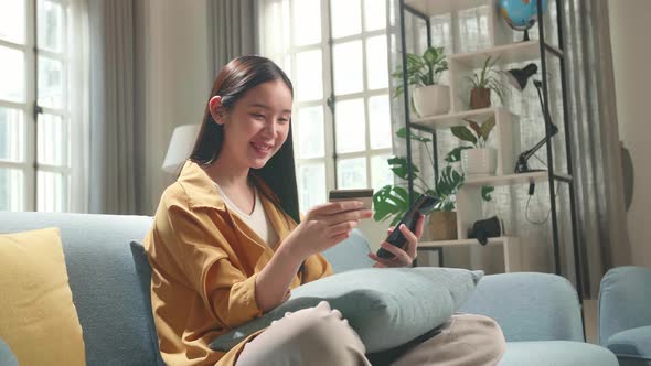 Asian Woman Sitting On Couch In Living Room Holding Credit Card And Using Smartphone Shopping Online