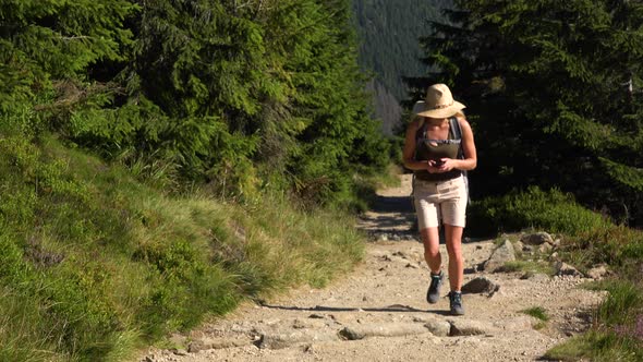 A Young Beautiful Woman Walks Up a Trail on a Hill with a Smartphone in Her Hands