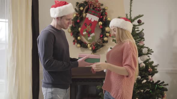 Young Caucasian Man in New Year Hat Giving Gift Box To Pretty Woman. Cute Girl Opening Present and