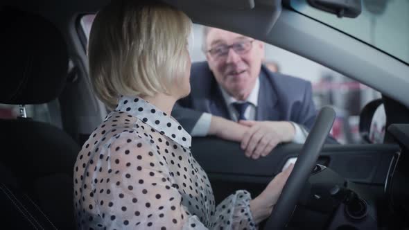 Side View of Beautiful Mature Caucasian Woman Sitting on Driver's Seat and Talking with Elderly