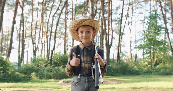 Boy in Straw Hat which Posing on Camera with Fishing Rods in the Green Park and Showing Sign Ok