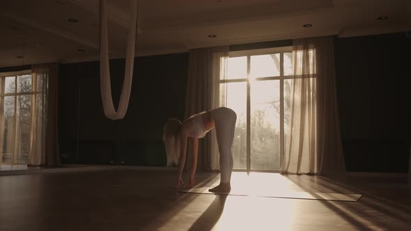 A Young Woman in White Sportswear is Stretching with a Large Hall with Large Windows in a Slowmotion