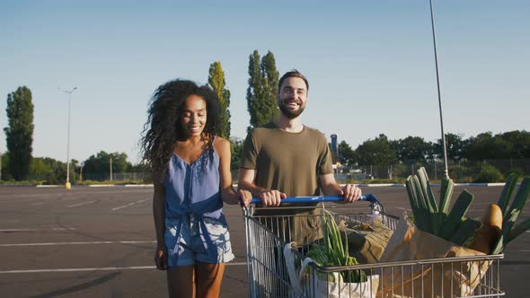 Young Multiracial Pair Laughing Walking By Empty Car Parking with Trolley of Groceries After